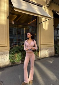 Jasnoróżowa bluzka na długi rękaw z zamkiem i rozkloszowane spodnie wykonane z mi�ękkiego materiału. Szarobrązowe botki za kostkę i okulary przeciwsłoneczne w ręce. Zielone liście w tle.