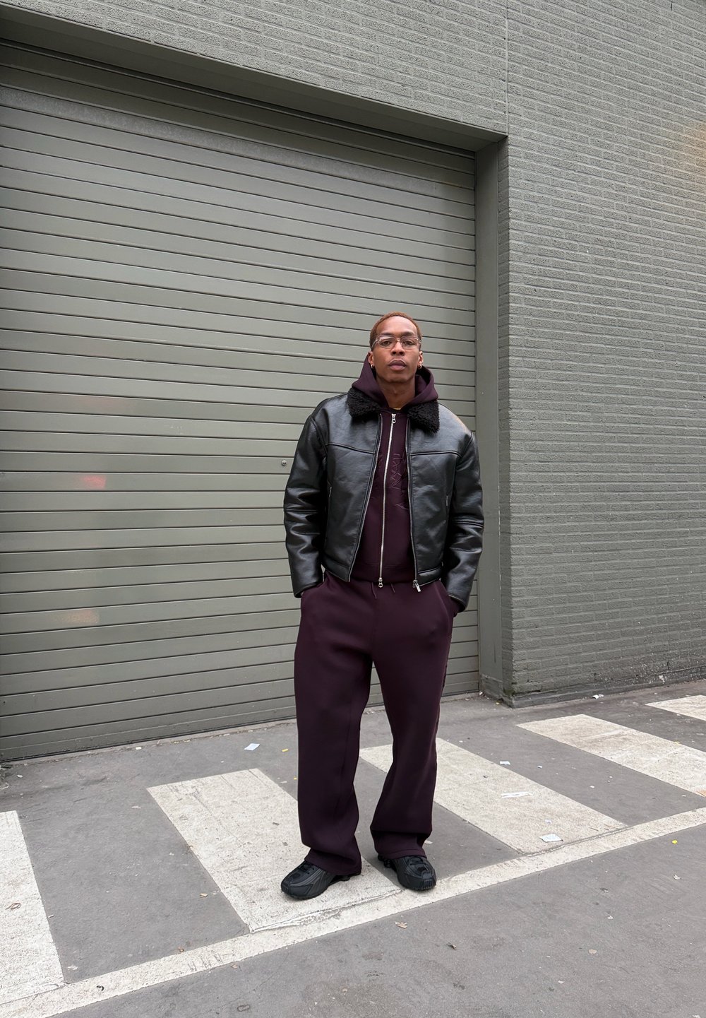 Veste bomber en cuir noir avec col en fourrure, sweat à capuche bordeaux et pantalon de survêtement assorti. Baskets noires, debout contre un mur gris.