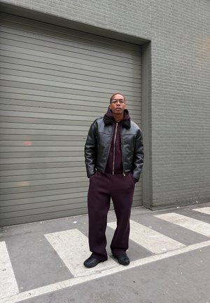 Cazadora bomber de cuero negro con cuello de piel, sudadera con capucha burdeos y pantalones deportivos a juego. Zapatillas negras, de pie contra una pared gris.