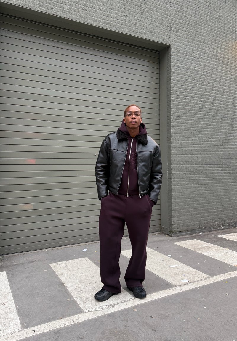 Veste aviateur en cuir noir avec col en fourrure, sweat à capuche bordeaux et pantalon de survêtement assorti. Baskets noires, debout contre un mur gris.