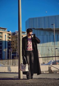 Personne portant un long manteau noir et une chemise rayée, tenant un sac à main blanc et buvant dans une tasse dans une rue urbaine ensoleillée.