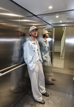 Hombre con pantalones deportivos grises, chaqueta vaquera y gorra blanca de pie en un ascensor de metal con un espejo que refleja su perfil lateral.