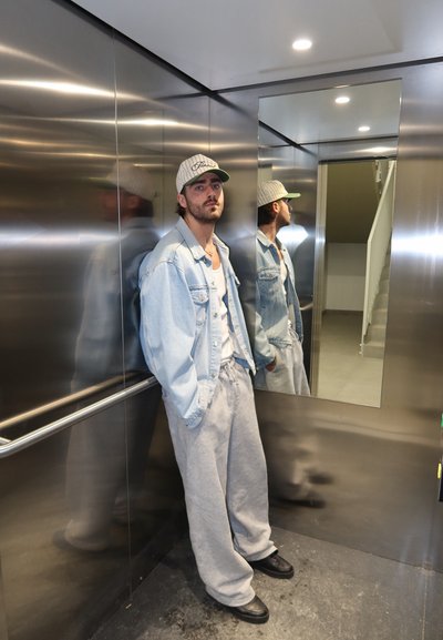 Hombre con pantalones deportivos grises, chaqueta vaquera y gorra blanca de pie en un ascensor de metal con un espejo que refleja su perfil lateral.