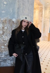 Femme aux longs cheveux bruns portant un béret blanc des New York Yankees, un manteau noir et tenant un sac noir devant un mur texturé usé.