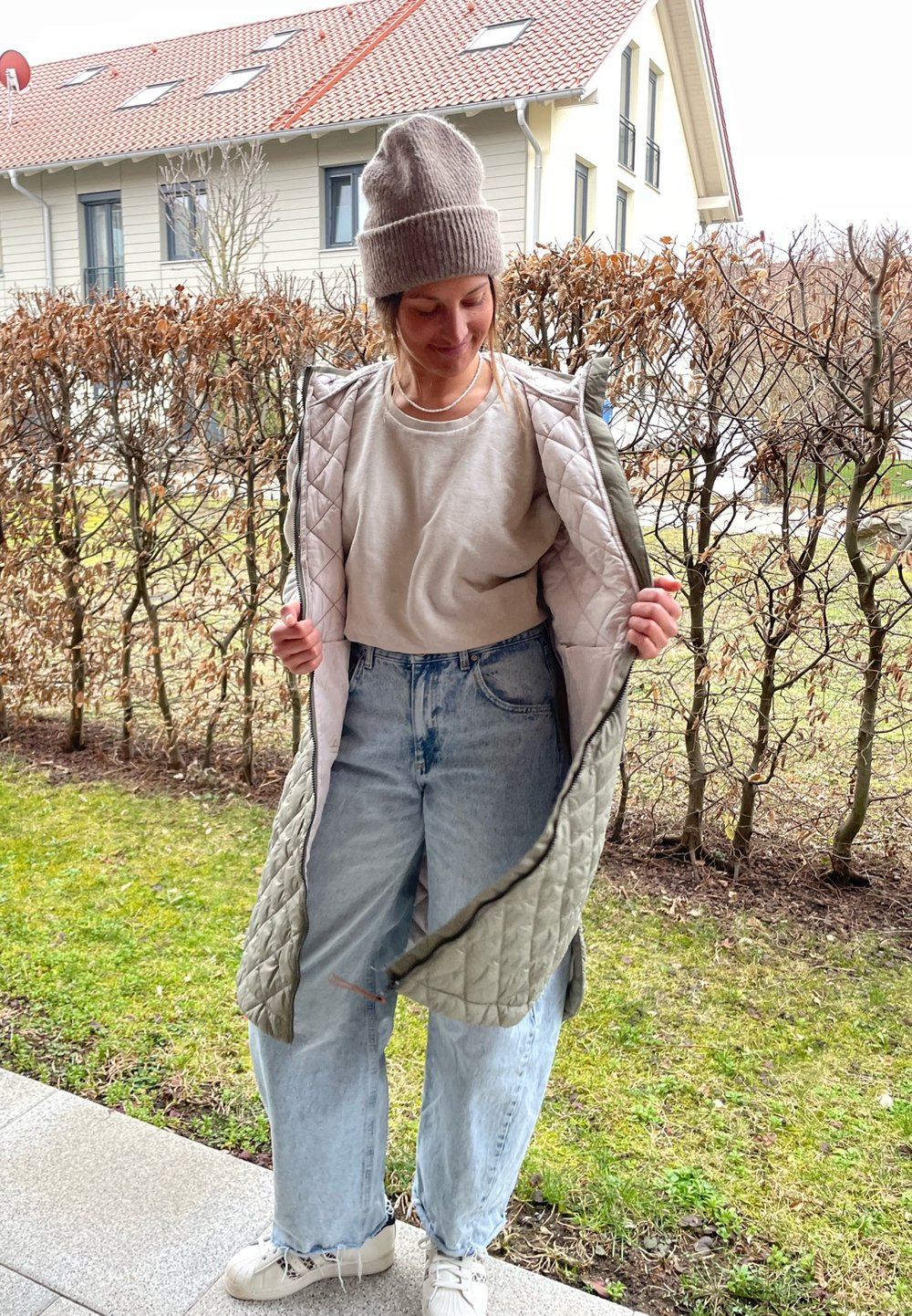 Κουβερτίνα μακρύ γιλέκο σε ελιά απόχρωση με ανοιχτό εσωτερικό, φορεμένο πάνω από μπεζ πουκάμισο και χαλαρά ανοιχτό μπλε τζιν. Λευκά παπούτσια ολοκληρώνουν την εμφάνιση.