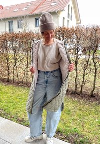 Κουβερτίνα μακρύ γιλέκο σε ελιά απόχρωση με ανοιχτό εσωτερικό, φορεμένο πάνω από μπεζ πουκάμισο και χαλαρά ανοιχτό μπλε τζιν. Λευκά παπούτσια ολοκληρώνουν την εμφάνιση.