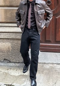 Brązowa skórzana kurtka z dwoma przednimi kieszeniami, czarna koszula z guzikami i brązowy krawat. Czarna para dżinsów i wypolerowane czarne buty dopełniają stylizację.