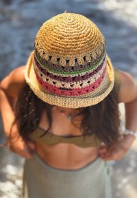 Άτομο με πολύχρωμο καπέλο κροσέ, μπικίνι τοπ σε ελαιώδη απόχρωση και φούστα σε ανοιχτό χρώμα, στέκεται σε υπαίθριο χώρο με θολό φόντο.