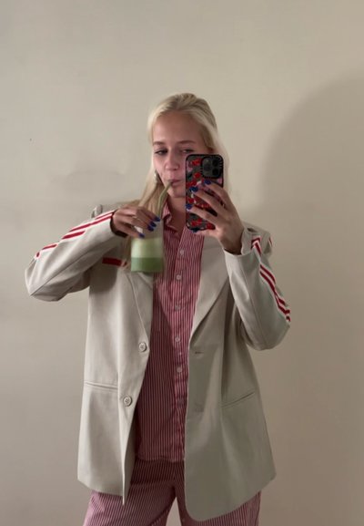 Blazer beige con acentos de rayas rojas sobre una camisa y pantalones de rayas rojas y blancas. Sosteniendo una bebida verde en un vaso.