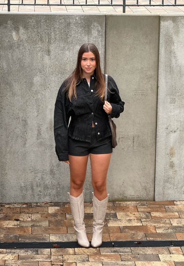 Mlada ženska stoji ob betonskem zidu, oblečena v črno jeans jakno, črne kratke hlače, bele kavbojske škornje in nosi torbico čez ramo.