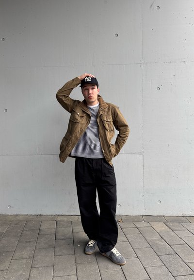 Joven con chaqueta marrón, camisa de rayas, pantalones negros y gorra negra, de pie sobre un pavimento de baldosas frente a una pared de hormigón gris.
