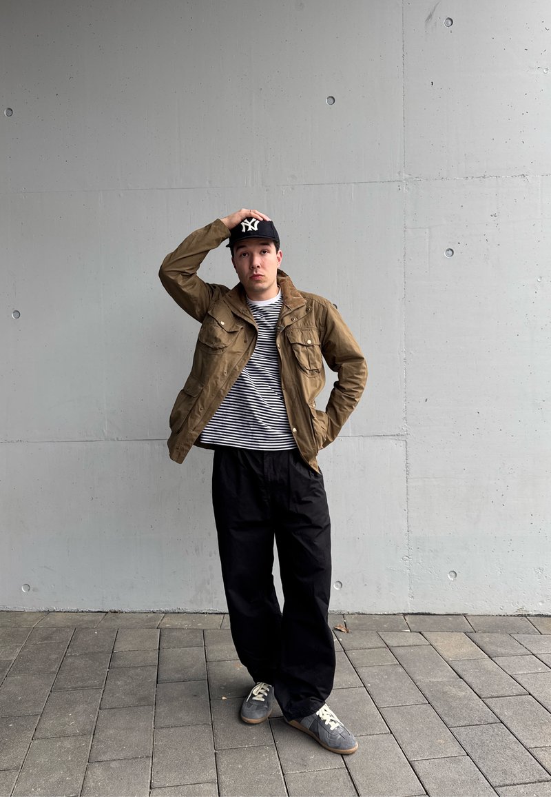 Jeune homme en veste marron, chemise rayée, pantalon noir et casquette noire, debout sur un pavé carrelé devant un mur en béton gris.