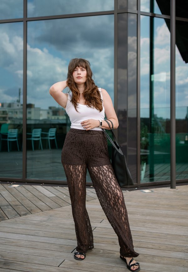 Vrouw met lang bruin haar die een wit tanktopje en zwarte kanten broek draagt, staat op een houten dek buiten een glazen gebouw en draagt een zwarte totebag.