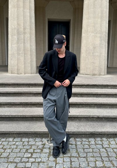 Blazer negro sobre una blusa negra, pantalones anchos grises y zapatos negros con calcetines blancos. Gorra negra con el logo de "LA". Fondo de piedras pavimentadas.