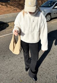 Femme portant une casquette beige et une veste polaire blanche, tenant un sac matelassé beige, marchant dans la rue près d'une voiture argentée garée.