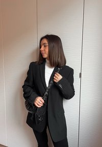 Jeune femme aux cheveux bruns mi-longs portant un blazer noir, un col roulé blanc, tenant une sangle de sac à main noire texturée contre un mur clair texturé.