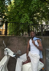 Kermanvärinen skootteri kromiyksityiskohtineen, asetettuna tekstuuripinnoitteiseen kiviseinään, taustalla vihreyttä ja henkilö rentoina vaatteissa.