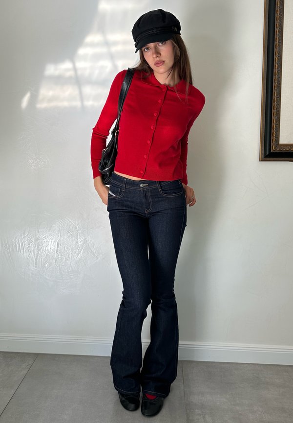 Joven mujer con una blusa roja abotonada, vaqueros oscuros acampanados, sombrero negro y zapatos negros, de pie en el interior contra una pared blanca.