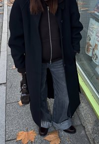 Črni dolg plašč čez temno kapuco z zadrgo, združen s širokimi sivimi jeans hlačami. Rjava usnjena torba in sijoči temni čevlji, ob katerih so padli listi.