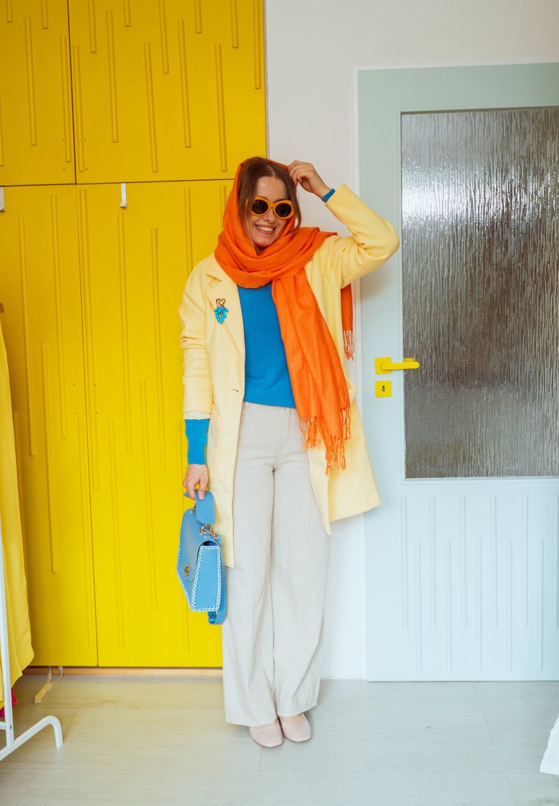 Femme portant une écharpe orange et des lunettes de soleil, un manteau jaune, un pull bleu, tenant un sac à main bleu, debout à côté de casiers jaunes et d'une porte.