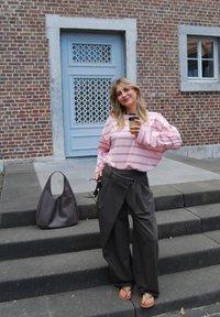 Helles rosa gestreiftes Langarmoberteil, dunkelblaue weiter Beine Hose, braune geflochtene Tasche, hält eine Tasse Kaffee, steht auf Steinstufen neben einer blauen Tür.