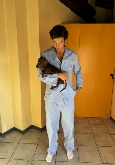 Conjunto de pijama a rayas azul claro de algodón, que incluye una camisa con botones y un bolsillo, y pantalones de corte holgado. Un pequeño perro marrón es sostenido en brazos.