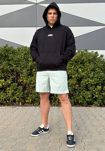 Joven con sudadera negra con capucha puesta, pantalones cortos claros y zapatillas negras, de pie sobre la acera con arbustos y una pared geométrica detrás.