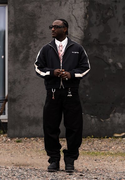 Hombre vestido con chaqueta negra con rayas blancas, corbata rosa, pantalones negros con llaveros, gafas de sol y anillos, de pie sobre grava junto a una pared de hormigón.
