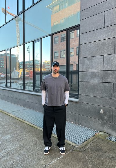 Pantalones anchos negros, camiseta gris de manga corta superpuesta sobre una camiseta blanca de manga larga, gorra negra y zapatillas negras con suelas blancas. Fondo urbano.