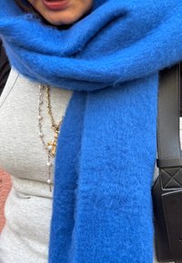 Écharpe en laine bleu roi avec une texture douce et duveteuse, enroulée autour du cou. Un haut gris côtelé et une sangle de sac noire sont partiellement visibles.