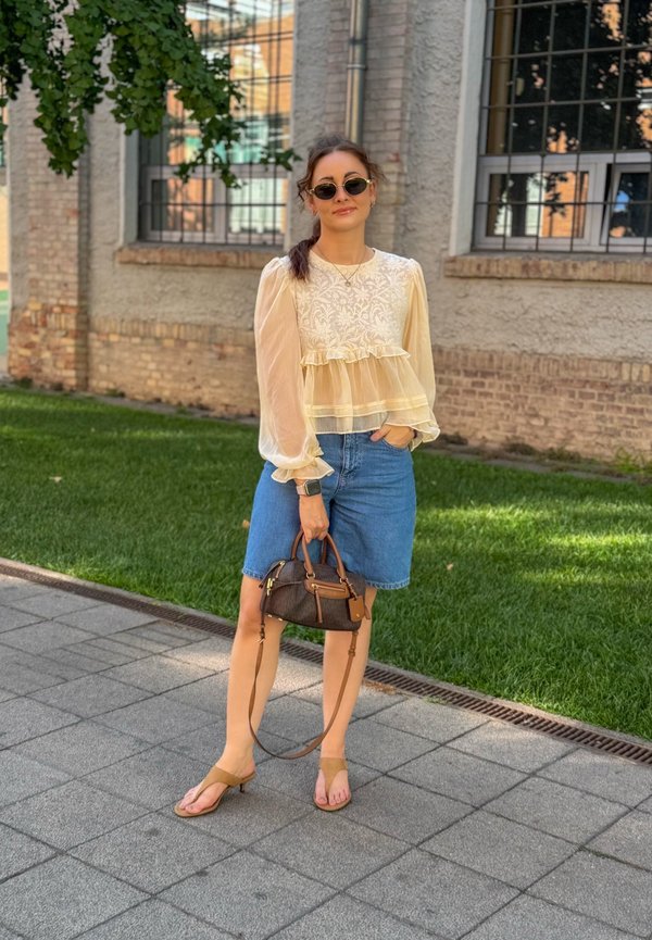 Femme portant un chemisier crème transparent et une jupe en jean, tenant un sac à main marron, debout sur le trottoir près de l'herbe et d'un bâtiment en briques avec des fenêtres grillagées.