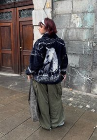 Veste en polaire noire avec un imprimé cheval gris, associée à un pantalon cargo vert ample. Sac gris avec des accents circulaires. Décor urbain en extérieur.