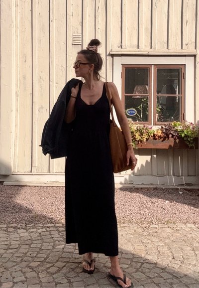 Vestido maxi negro sin mangas, corte holgado, sandalias planas, bolsa de mano marrón, sosteniendo una chaqueta negra, de pie en un camino de piedras cerca de un edificio de madera.