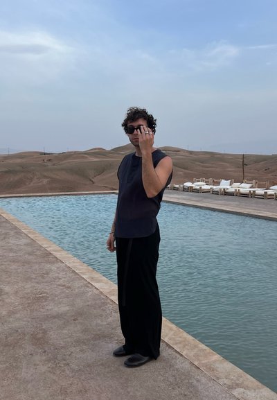 Top negro sin mangas, pantalones negros holgados, gafas de sol oscuras; de pie junto a una piscina turquesa con un paisaje arenoso de fondo.