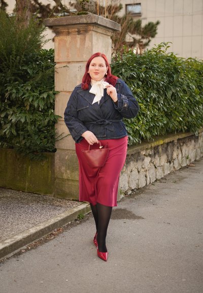 Mujer de cabello rojo con chaqueta azul marino, bufanda blanca, falda roja y zapatos rojos, caminando por una acera junto a vegetación y un muro de piedra.
