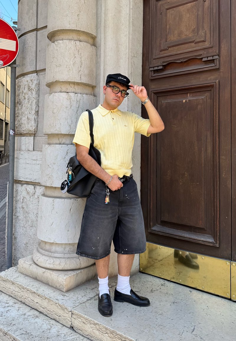 Mand iført gul polo shirt, sort kasket, briller og sorte shorts står ved en stenkolonne og en stor trædør og justerer sin kasket.