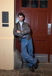 Ung man med mustasch iklädd läderjacka, bandana, rutig skjorta och jeans lutande mot en röd dörr på natten på en stenläggning.