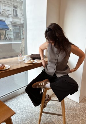 Mujer con cabello ondulado, gafas de sol en la cabeza, chaleco abotonado, pantalones negros y zapatillas, sentada en un taburete de madera junto a una mesa junto a la ventana con pasteles y café.