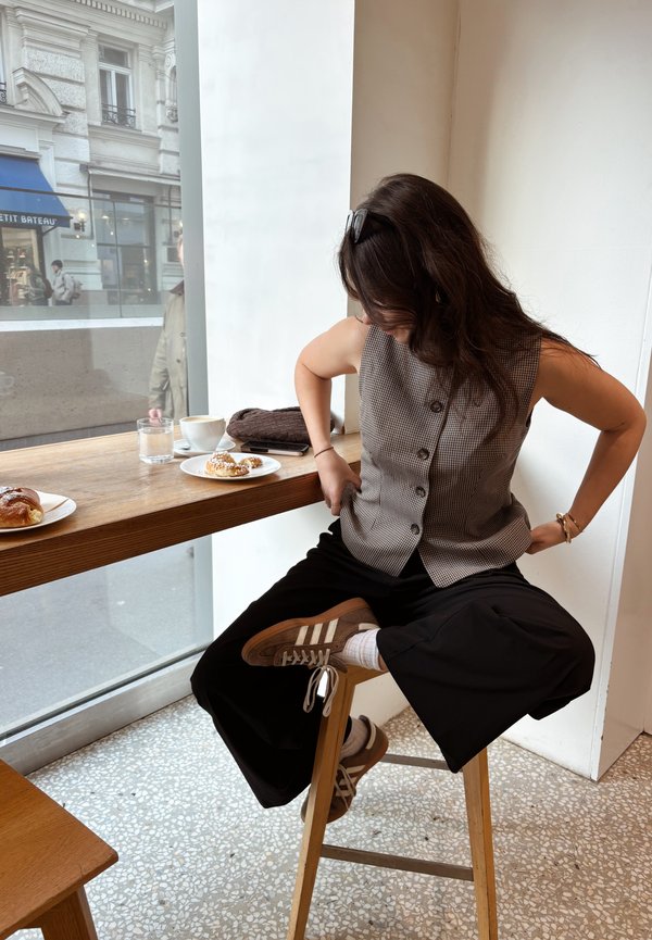Mujer con cabello ondulado, gafas de sol en la cabeza, chaleco abotonado, pantalones negros y zapatillas, sentada en un taburete de madera junto a una mesa junto a la ventana con pasteles y café.