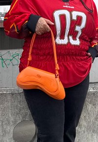 Oranžová lakovaná kožená kabelka s zakriveným, úzkym tvarom a krátkym remienkom, držená osobou oblečenou v červenom drese a čiernych džínsoch.