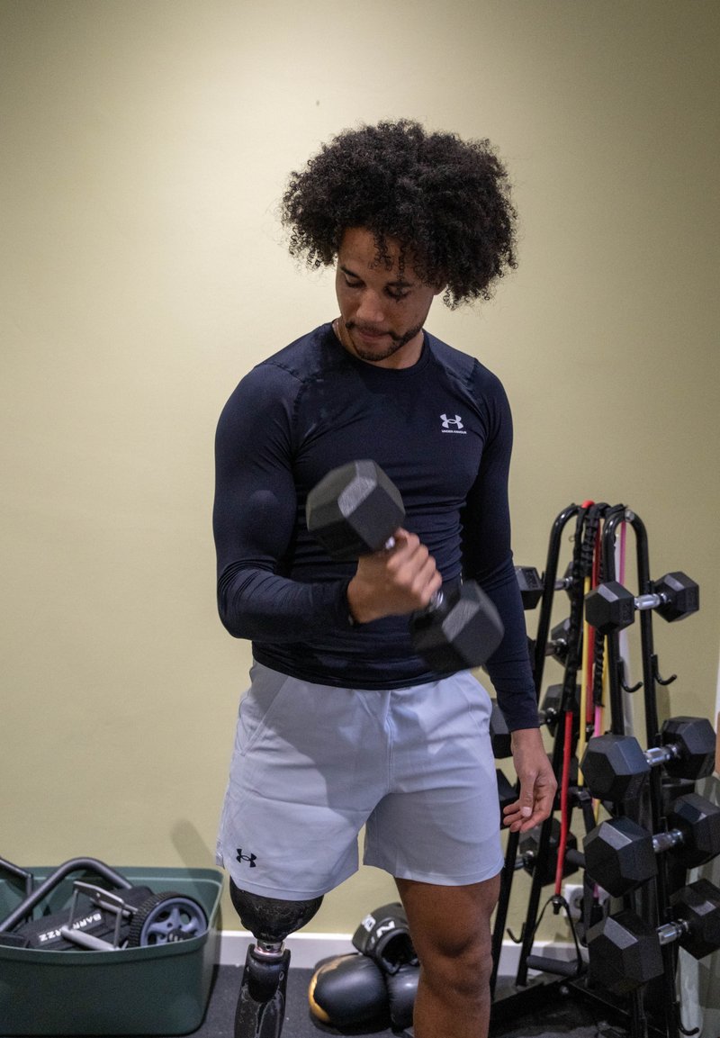 Camiseta preta de manga comprida para desporto, calções cinzentos, segurando um haltere hexagonal preto. Perna protética visível e equipamento de treino ao fundo.