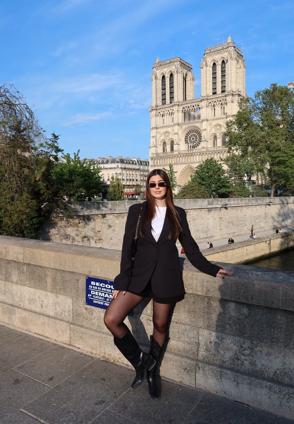 Joven mujer que lleva una chaqueta negra, pantalones cortos y botas apoyada en la barandilla de piedra del puente con la catedral de Notre-Dame al fondo en un día soleado.