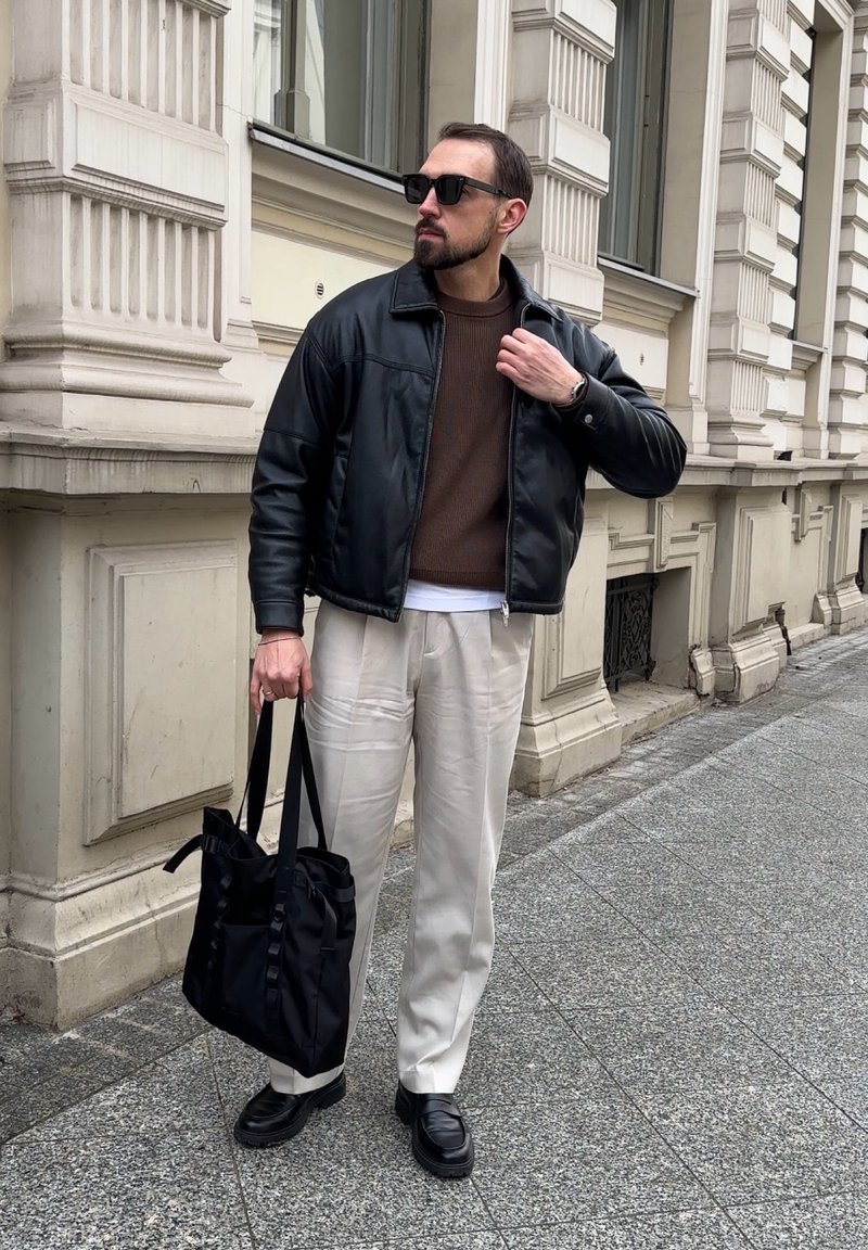 Homme portant une veste en cuir noire, un pull marron, un pantalon beige, des mocassins noirs, des lunettes de soleil, portant un sac fourre-tout noir sur un trottoir en ville.