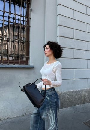 Mujer con cabello rizado, lleva una camiseta blanca de manga larga y vaqueros anchos, lleva un bolso negro y camina junto a un edificio de piedra con una ventana con rejas.