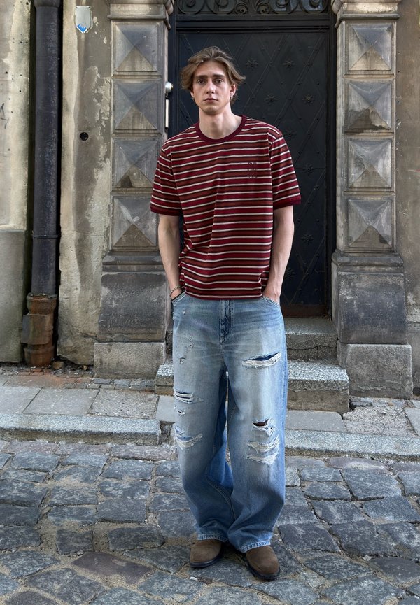 Ung mann med middels langt hår iført en rødstripet T-skjorte og revne, vide jeans står på en brosteinsgate foran en mørk dør.