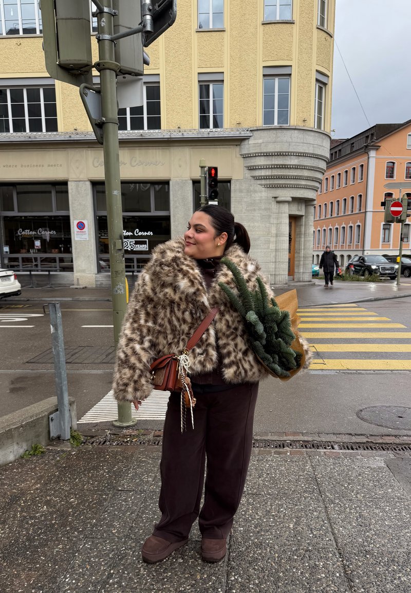 Bruin-wit gevlekte nepbontjas, loszittende donkerbruine broek, kleine bruine schoudertas en vasthouden van groen blad in bruin papier.