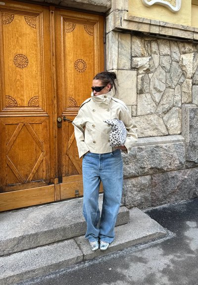 Mujer que lleva una chaqueta cruzada beige y jeans azules, sosteniendo un bolso con manchas, de pie en unos escalones de piedra junto a una puerta de madera ornamentada y una pared de piedra.