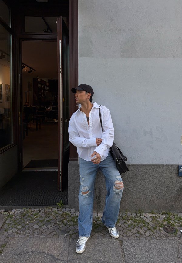 Junger Mann in weißem Hemd, zerrissenen Jeans und silbernen Sneakers lehnt an einer grauen Wand neben einer offenen Türöffnung.