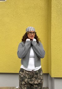 Femme aux cheveux bouclés, portant de grandes lunettes, un foulard à motifs, un pull gris zippé, une chemise blanche et un pantalon camouflage, debout contre un mur jaune.