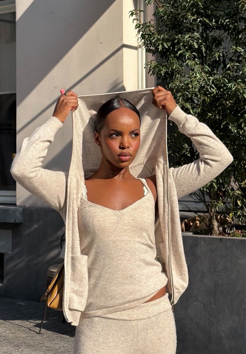 Mujer con top y falda de punto beige ajusta la capucha a juego al aire libre bajo la luz del sol, con vegetación y un edificio al fondo.